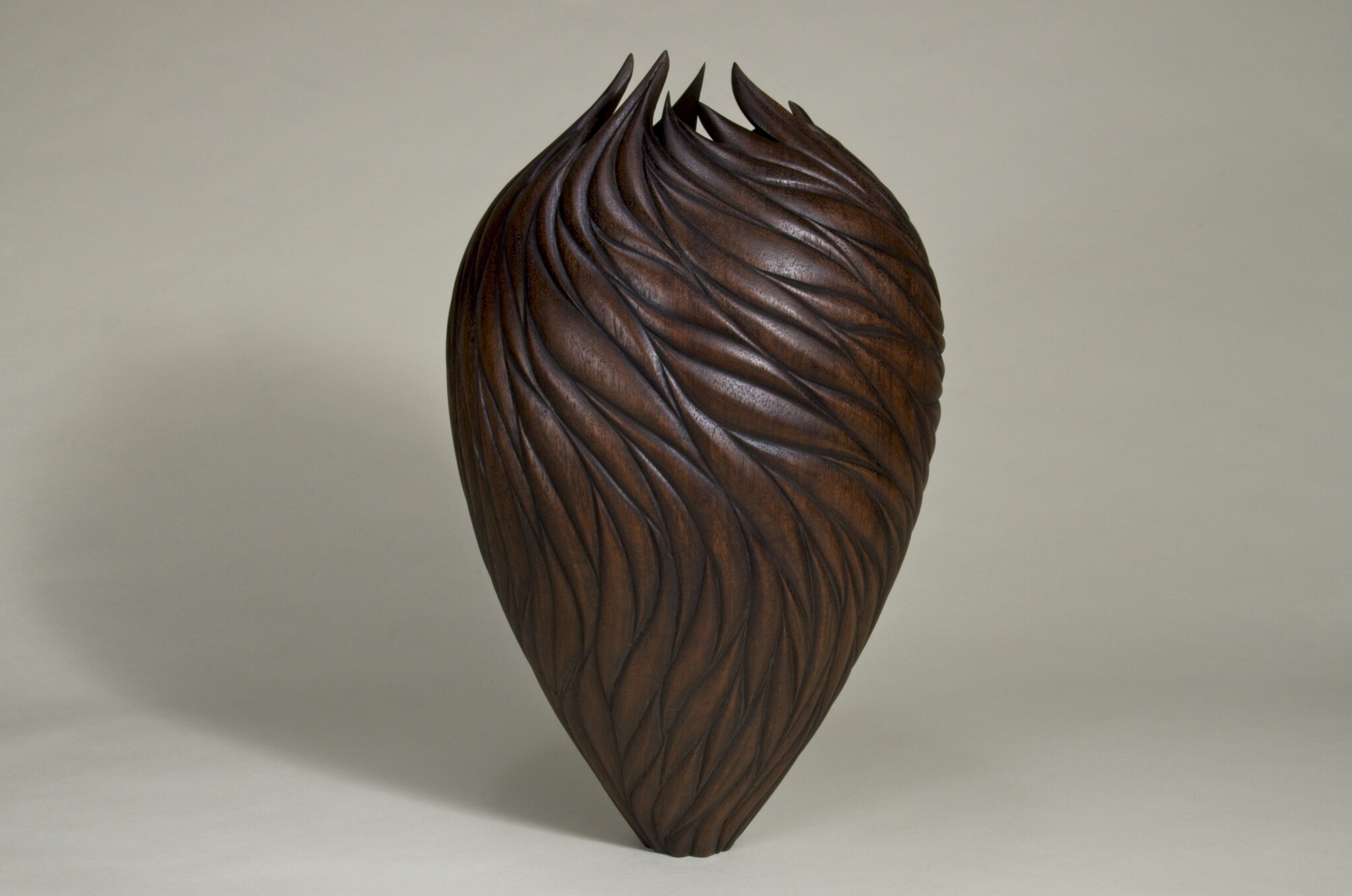 Dark brown wooden vase, shaped like an upside down pair. There is a waved pattern carved into the wood from the base to the top. The wood sticks out at in pieces around the top. 