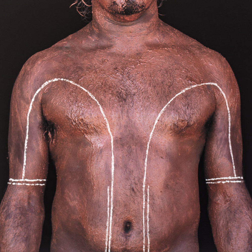 The torso of an Indigenous man against a black background. The man has lines painted up his torso and curved down his upper arm. The line ends in two straight lines around his elbow.