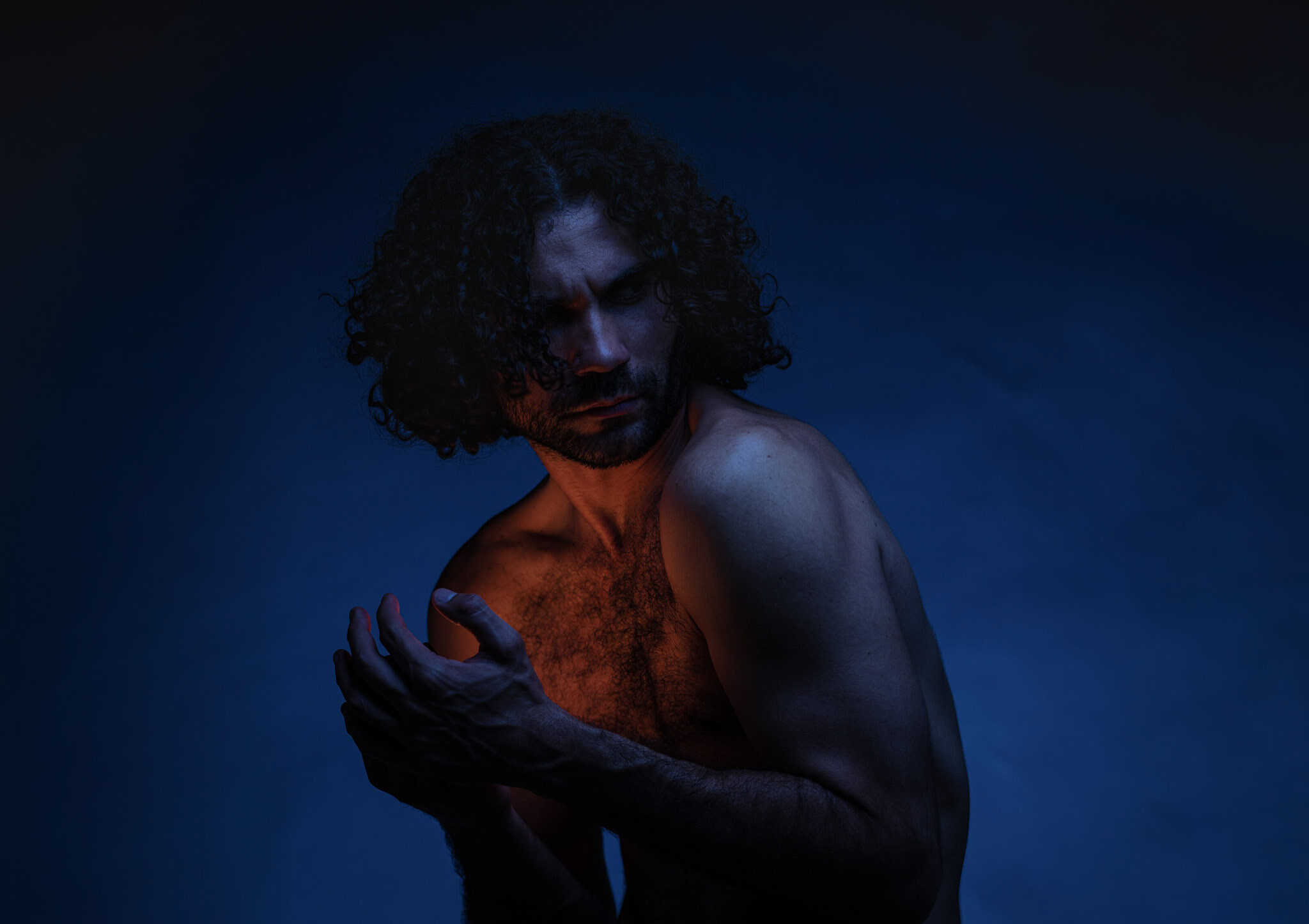 An Indigenous man with curly hair around his face bends over with his hands cupped in front of him. He looks to left over his shoulder. The background is a dark blue and an orange light shines from the inside of the man's hands onto his torso.