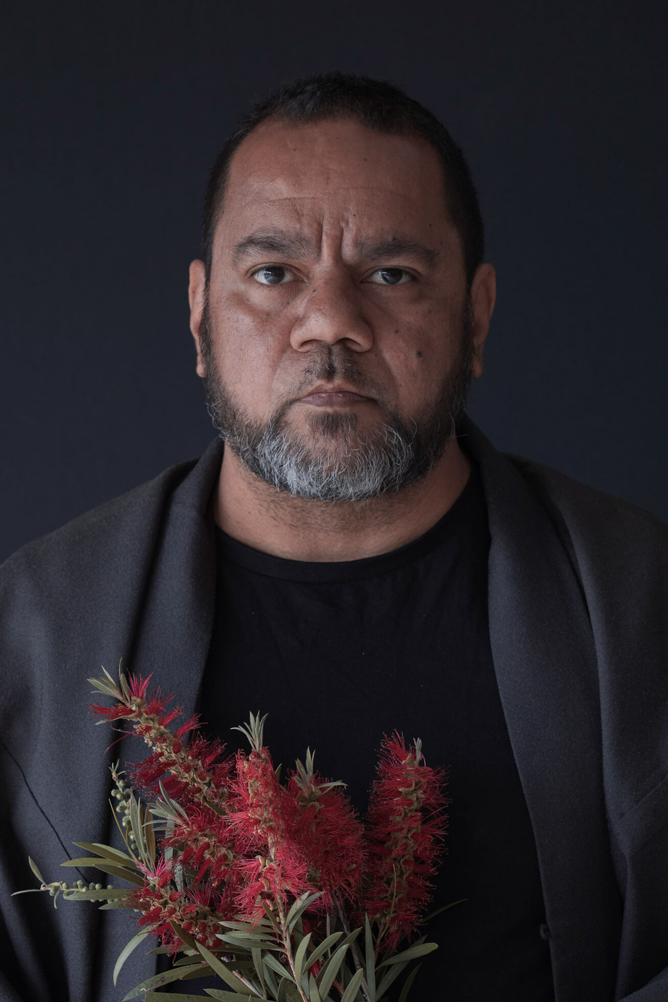 Michael is an Indigenous man with short hair and a short greying beard. He wears dark clothes, stands against a dark background and does not smile. In his hands is a bouquet of red bottlebrush flowers.