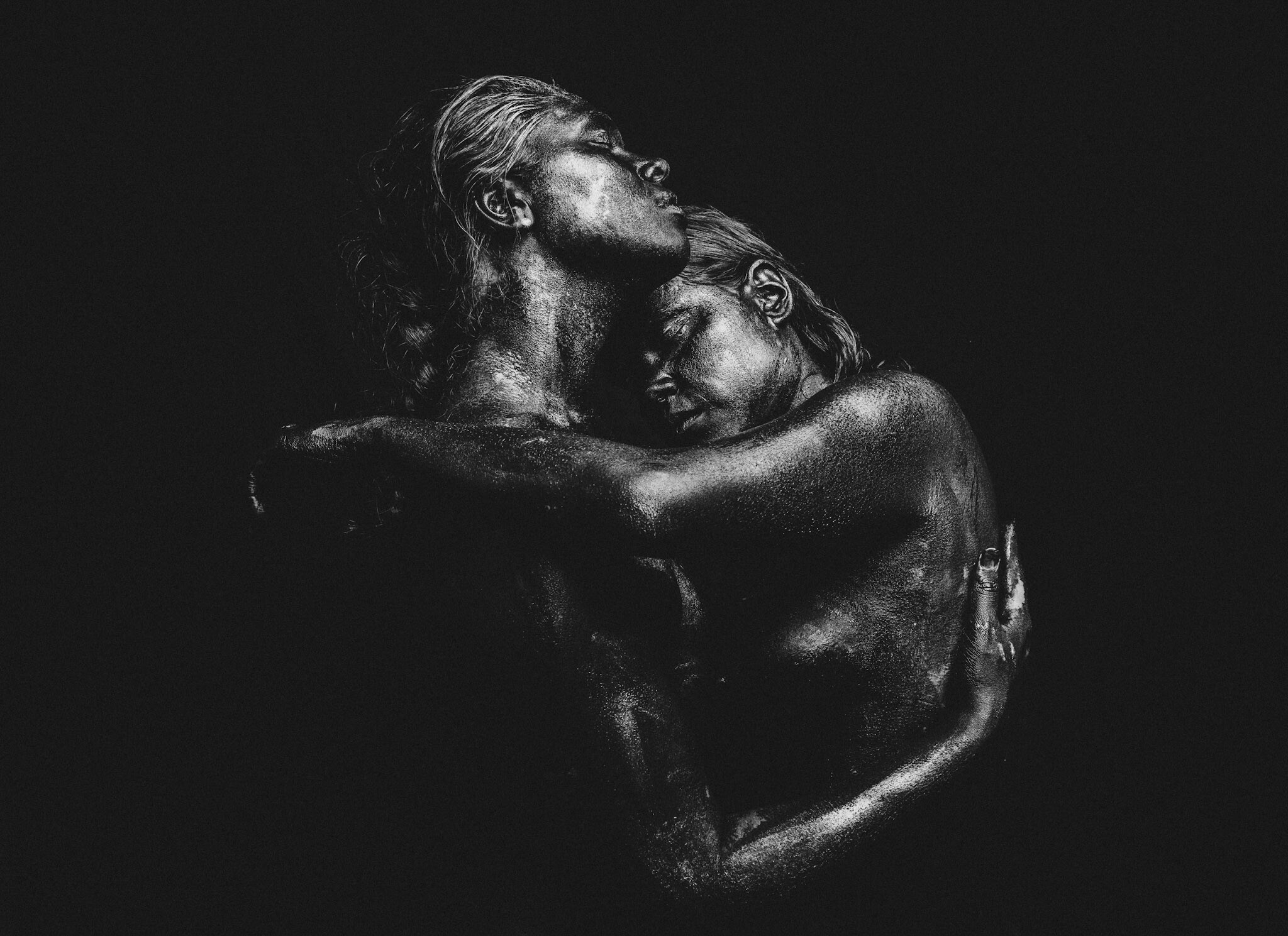 Two Indigenous women hugging with their eyes closed in close embrace. Image is very dark in black and white with a black background.