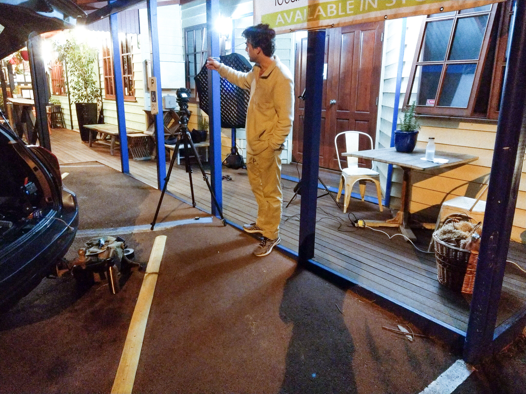 A young man stands on the front porch of a wel-lit film set pointing to the left.