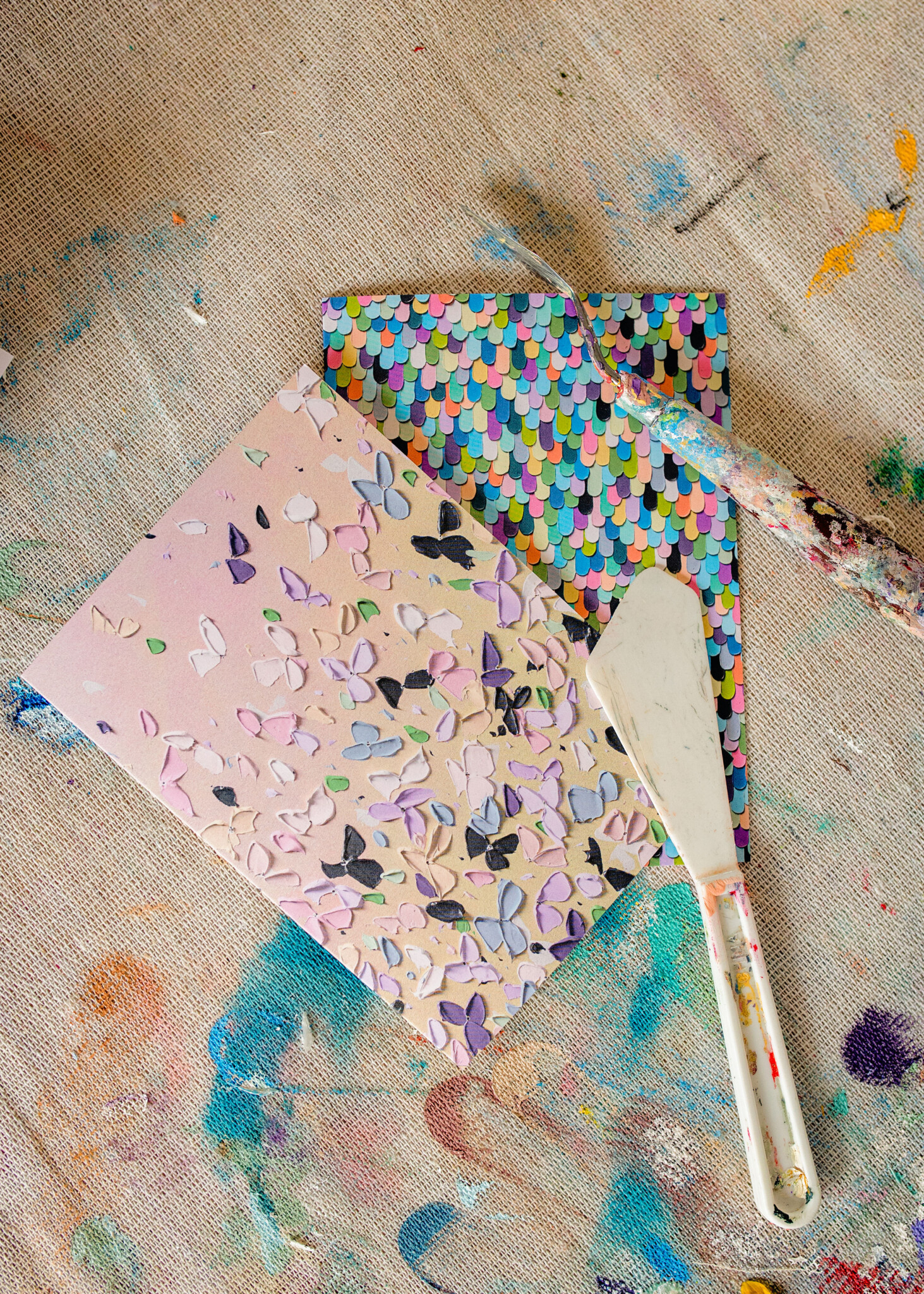 Two colourful and floral butterfly inspired artworks on top of each other, painting supplies are nearby.
