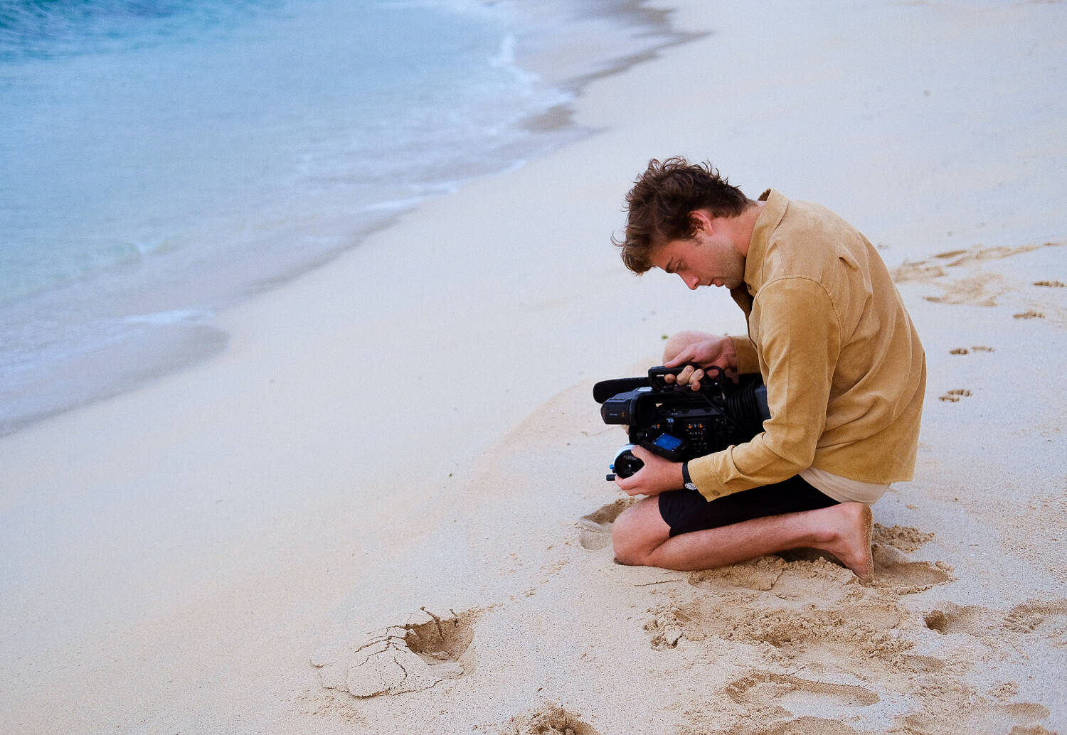 A young man sit on the beach filming the water.