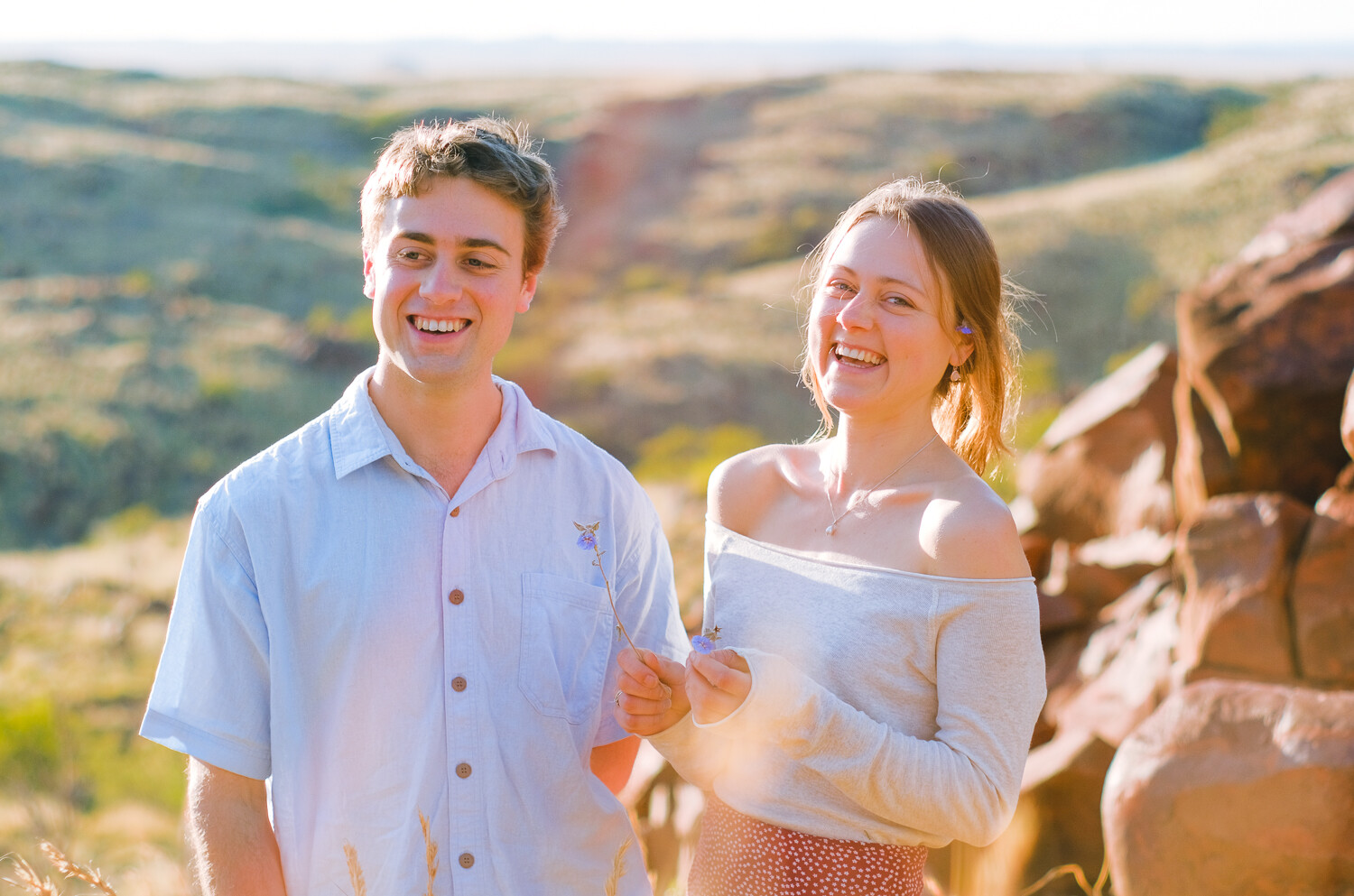 A young man standing next to a young woman. The young man smiles and looks away from camera wearing a white button down shirt. The youn woman smiles and looks at camera wearing an off-the-shoulder white shirt.