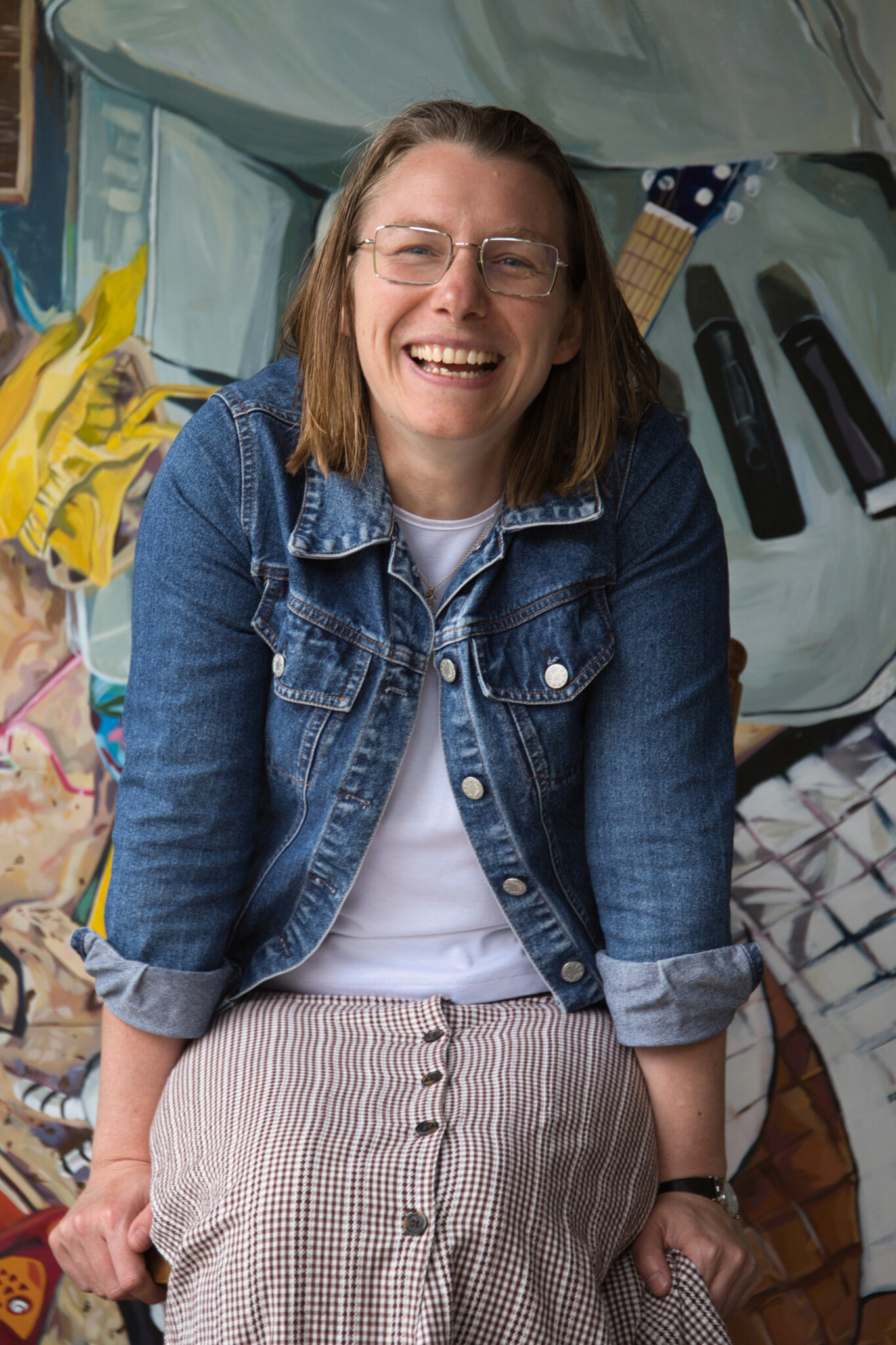 Elisha McGuckin sits and smiles at the camera. She has shoulder-length brown hair and wears clear-framed glasses. Her outfit is a white shirt, denim jacket and checkered skirt.