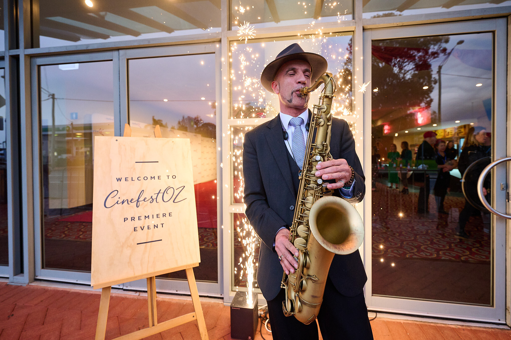 Man plays saxophone while sparks fly behind him, people lined up inside, a sign reads "WELCOME TO CINEFEST OZ"