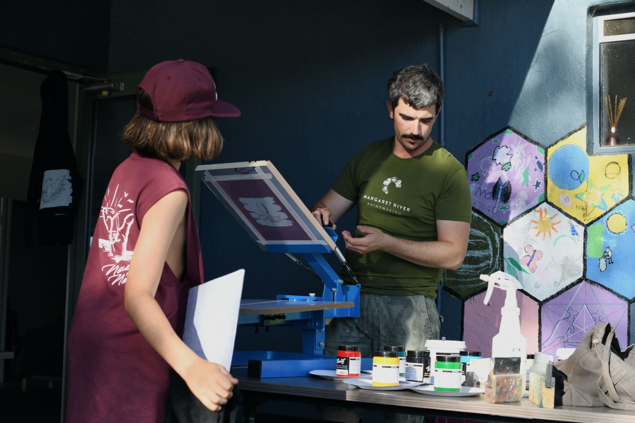 Francesco in a green T-Shirt reading 'Margaret River Printmaking' screenprints a design for a young person wearing a cap and tank top.