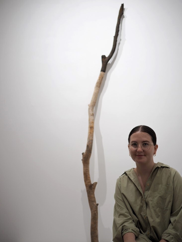 Rose wears a long khaki shirt and sits next to an artwork of a long upright tree branch.