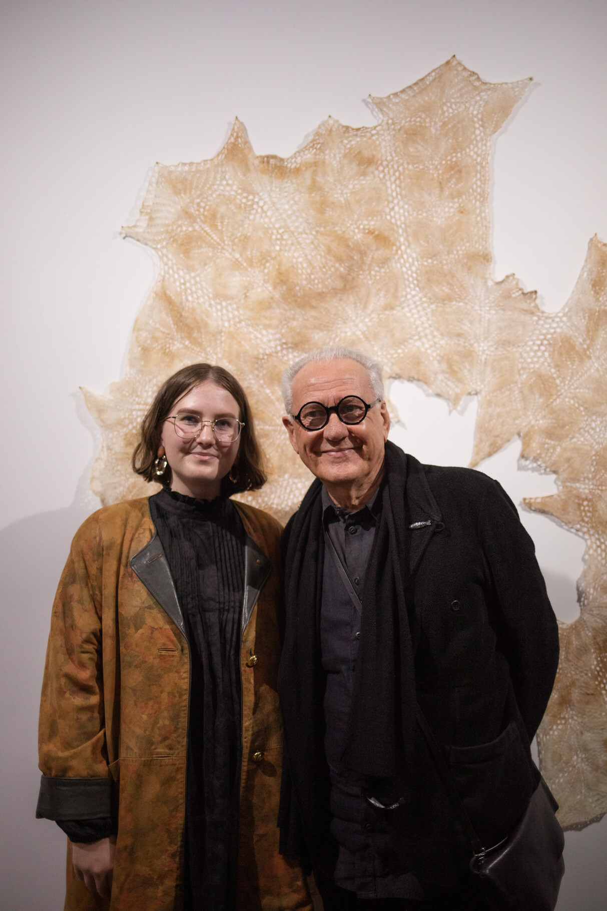 Rose wears a long brown jacket and clear glasses. She stands next to Ted Snell who wears a black jacket, black scarf and thick rimmed black round glasses.