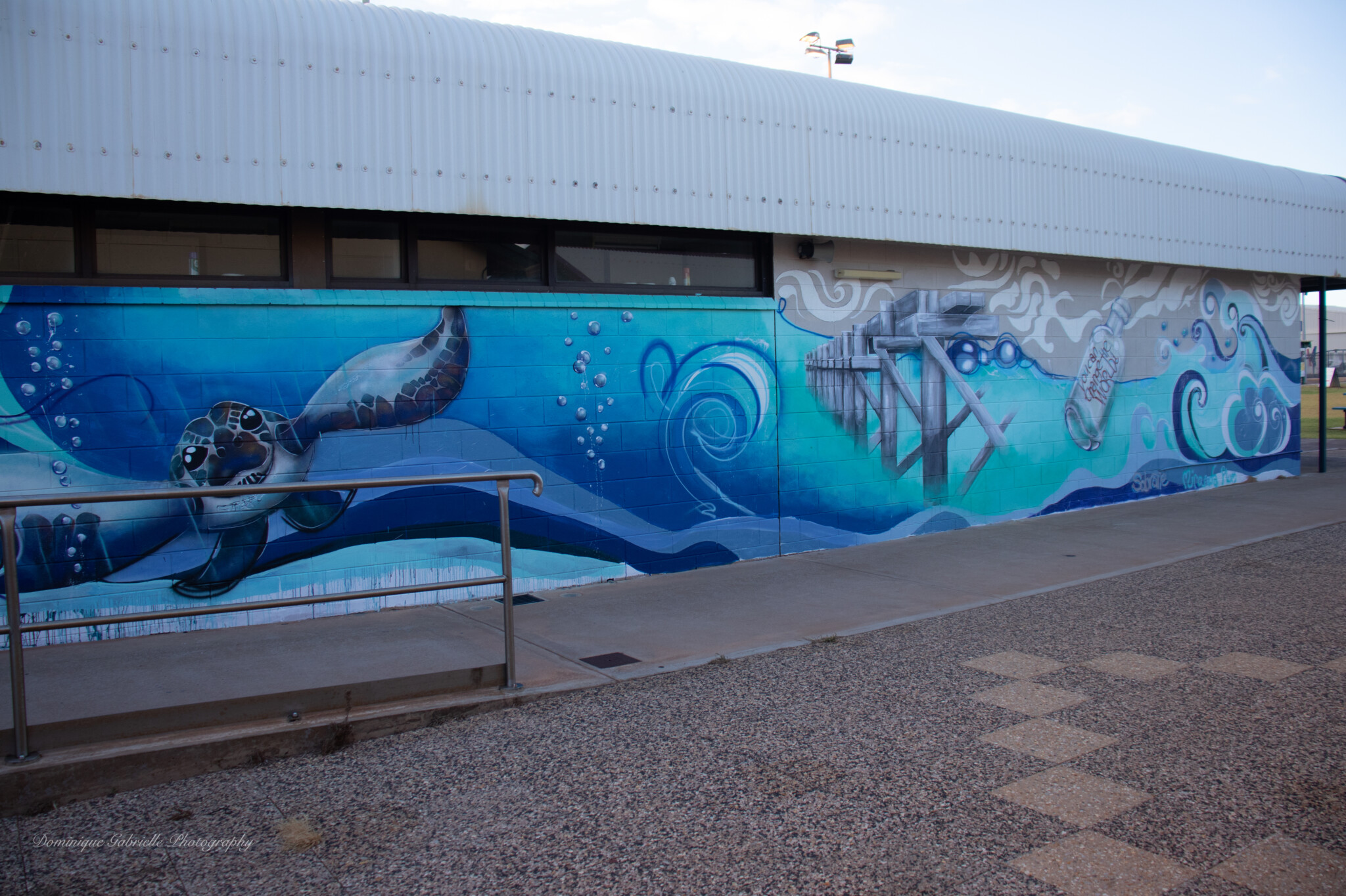 Mural of the ocean with sea animals, a message in a bottle and a pier on the side of a building.