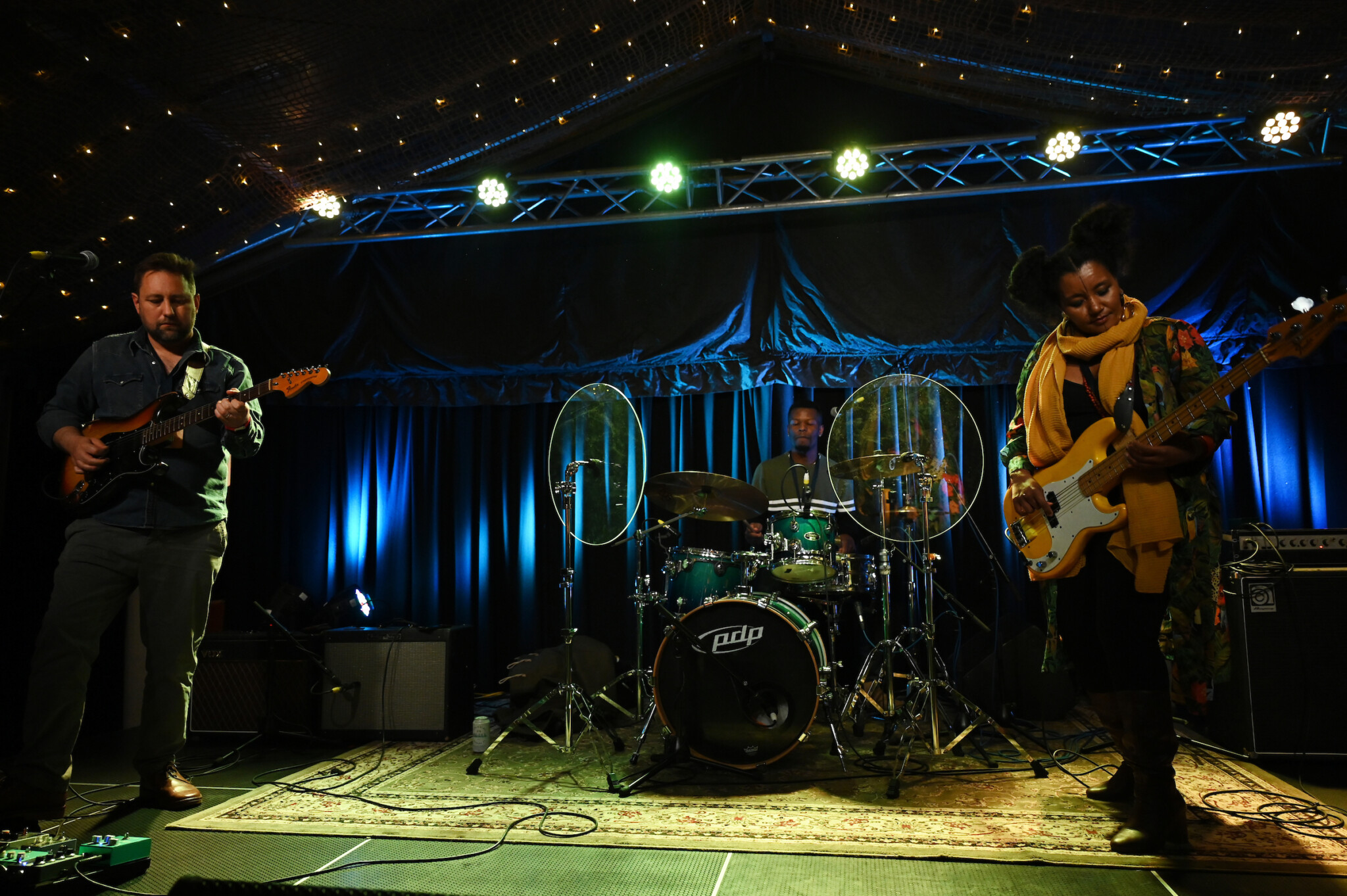 Live band performance with guitar, bass, and drums on stage lit by green and blue lights; string lights and sound shields visible.