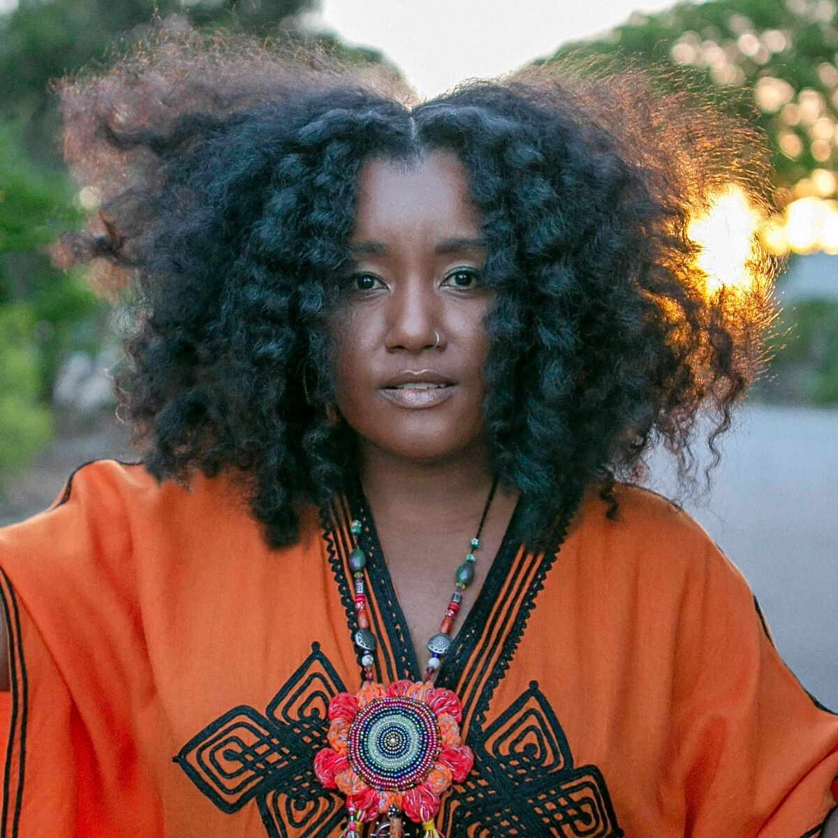 Person with curly hair in orange patterned garment and beaded necklace stands outdoors with greenery and setting sun.