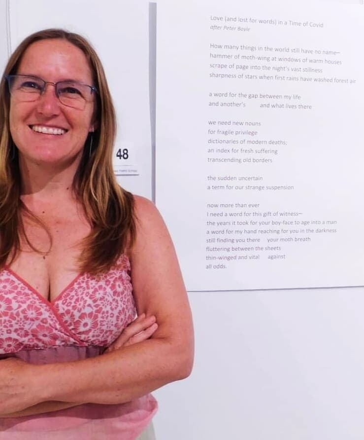 Renee wears a pink patterned top and stands smiling with her arms crossed in front of her printed poem on a gallery wall.