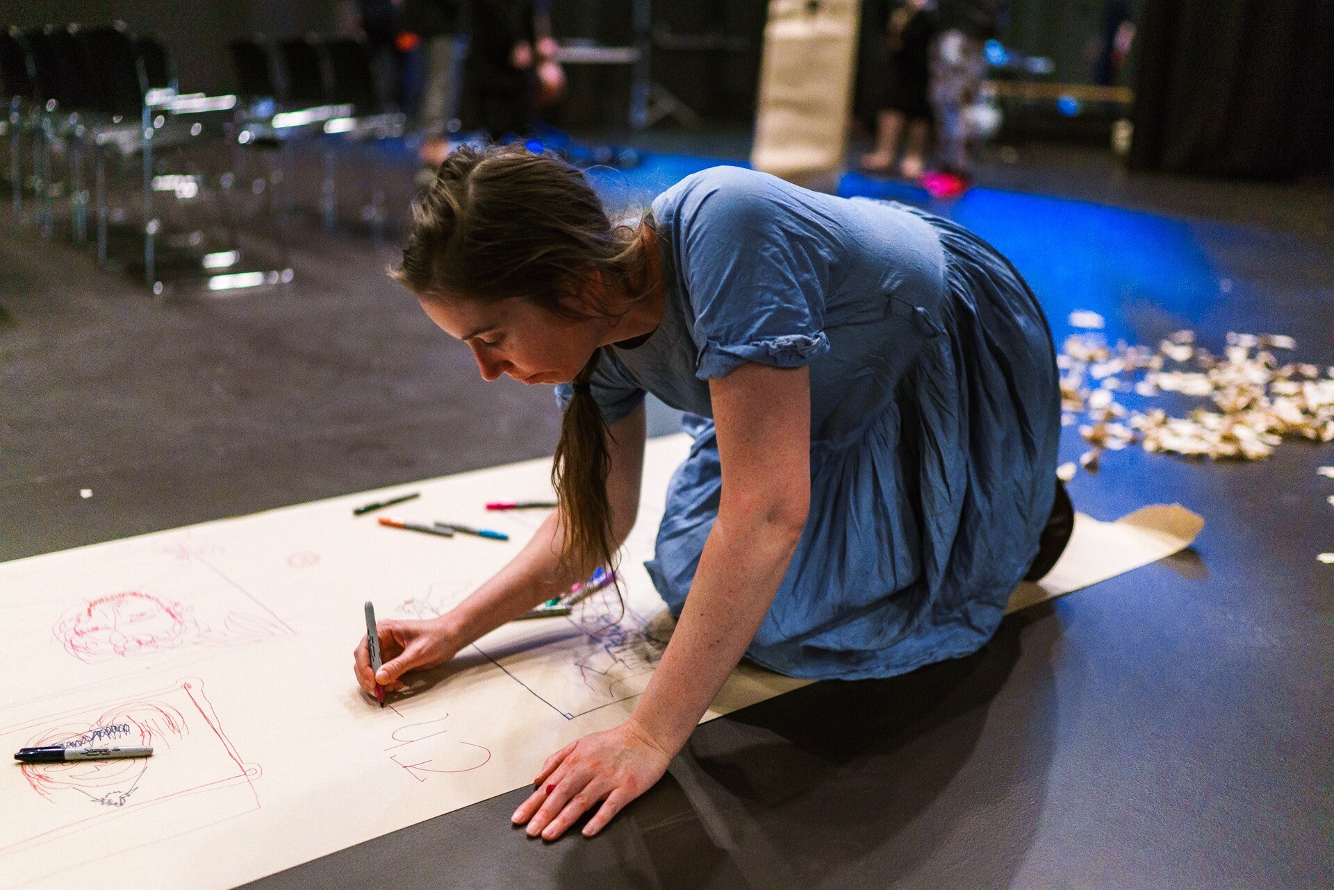 Chloe wears a denim dress and kneels on the floor drawing on a large piece of butchers papers.