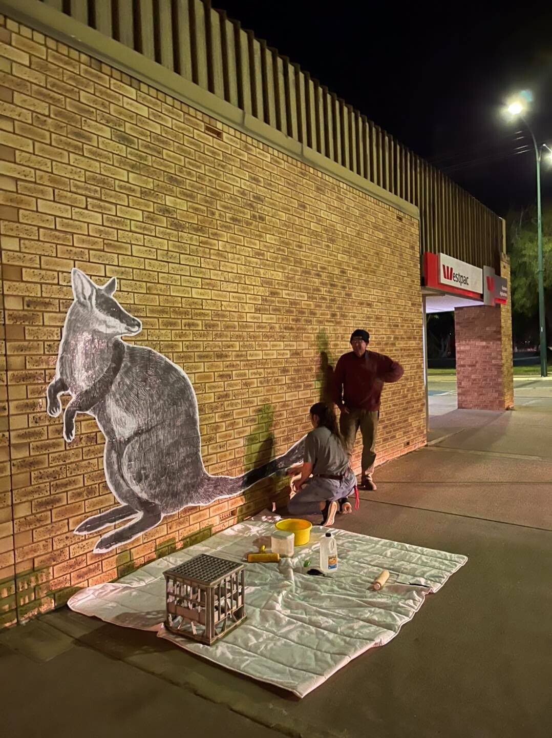 Two people pasting an image of a wallaby on a large brick wall.