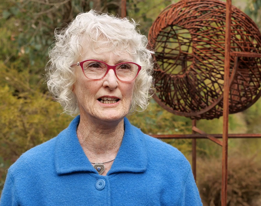 Person with white curly hair in blue jacket stands outdoors near rust-colored spherical metal sculpture.