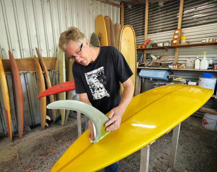 Man in shed fixing a yellow surfboard fin, with surfboards in the background.