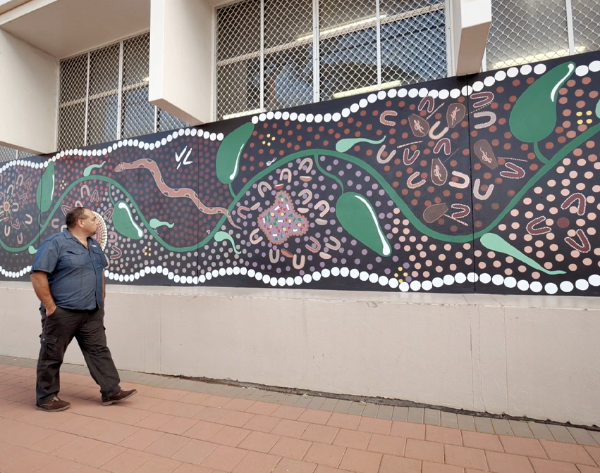 Person walks past abstract mural with green leaves, white dots, and brown-pink shapes on dark background with bright accents.