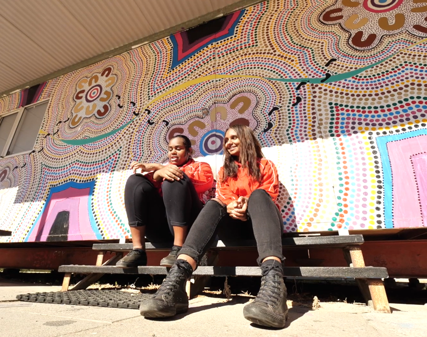 Two people in orange tops and black pants sit on black steps before wall of colorful dot art with flowers and abstract patterns.