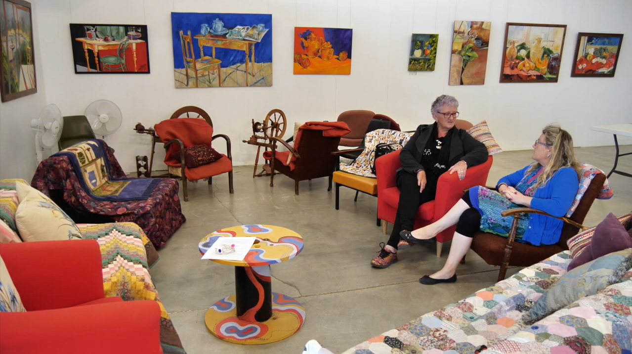 Art gallery with colorful paintings, concrete floor, fans, and two people seated among armchairs, a sofa, and a vibrant table.