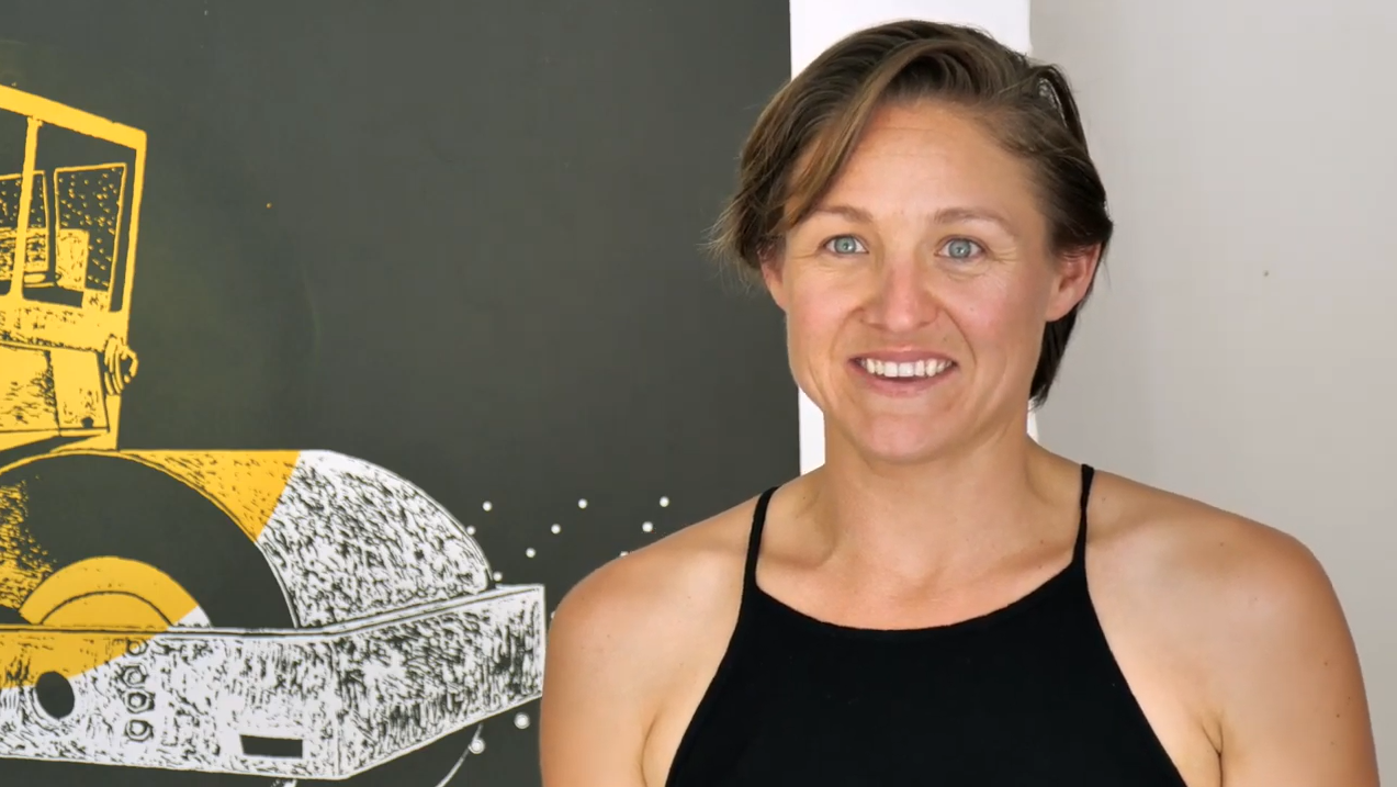 Woman with short brown hair smiles off camera, she wears a black singlet top and there is artwork of a drum roller behind her