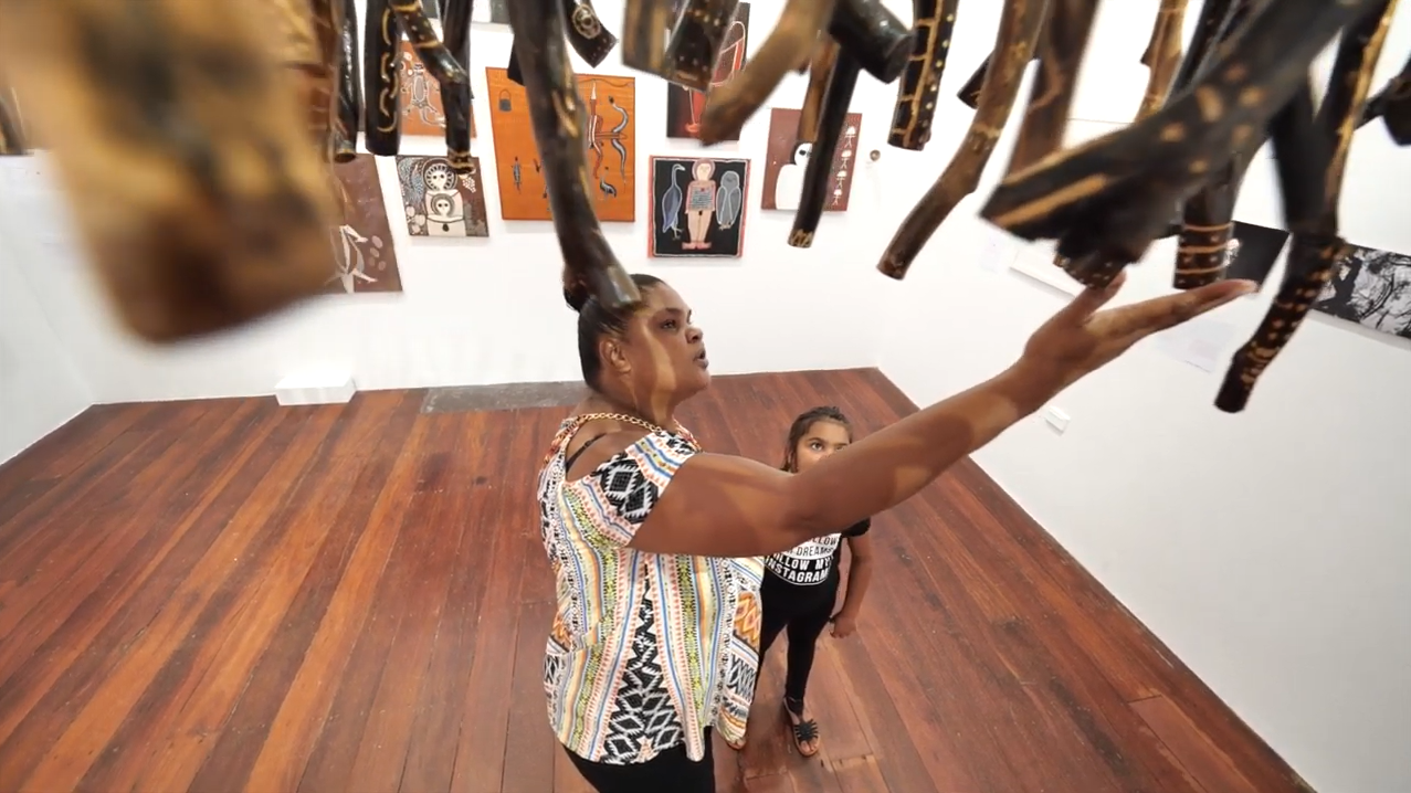 Two people in an art gallery with wooden floors; one raises an arm near wall art and ceiling-hung objects.