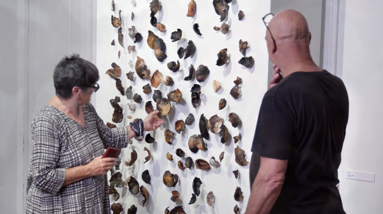 Two people stand before a white wall covered in small, irregular objects; one gestures with a red item while the other observes.