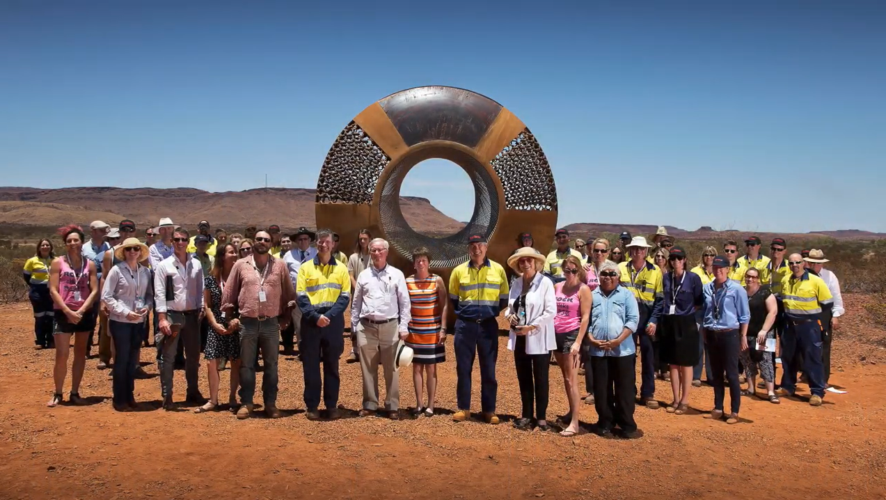 Group of people stand before a large metal sculpture with a central hole in a desert-like setting; mix of casual and work attire.