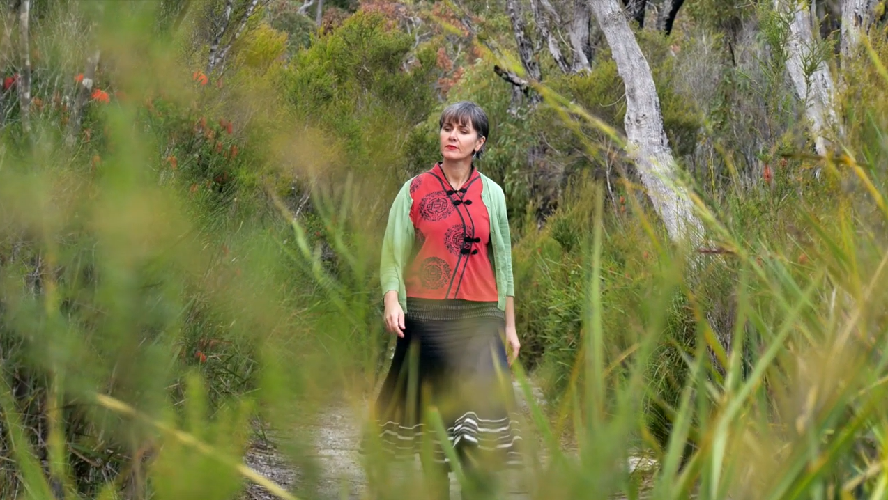 Woman wearing a red shirt, black skirt and green cardigan walks along a bush track surrounded by greenery.