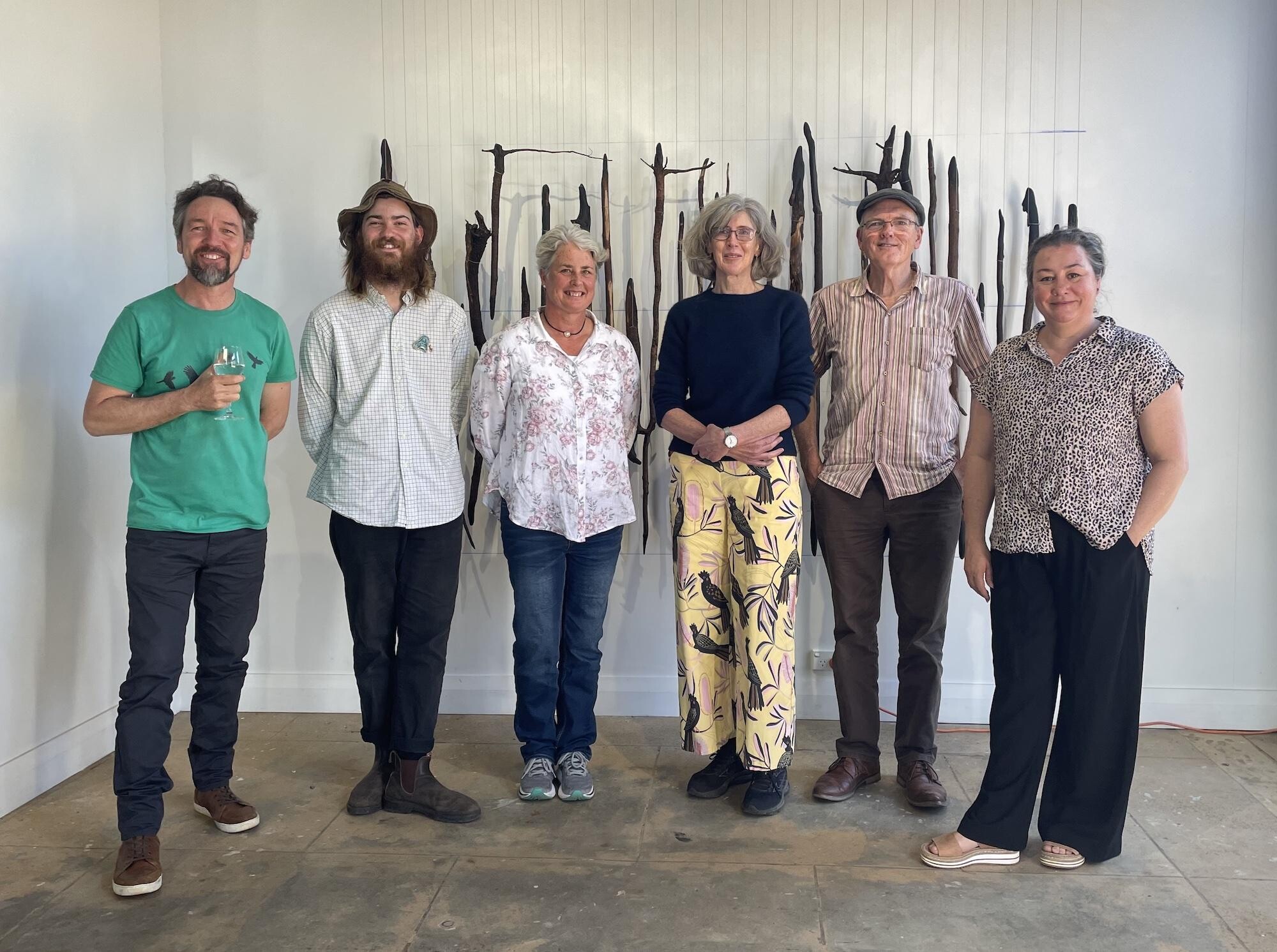 Six people stand in row indoors. Behind them, abstract wall installation of dark vertical sticks or branches.
