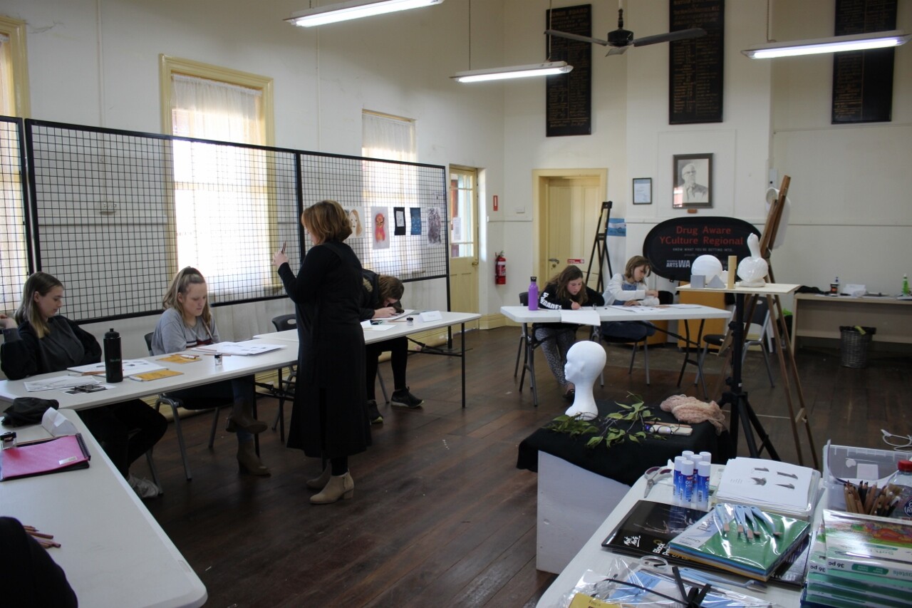 Art supplied are seen in the foreground, there are teenagers at tables doing artwork. A woman stands on the other side of the tables, teaching.