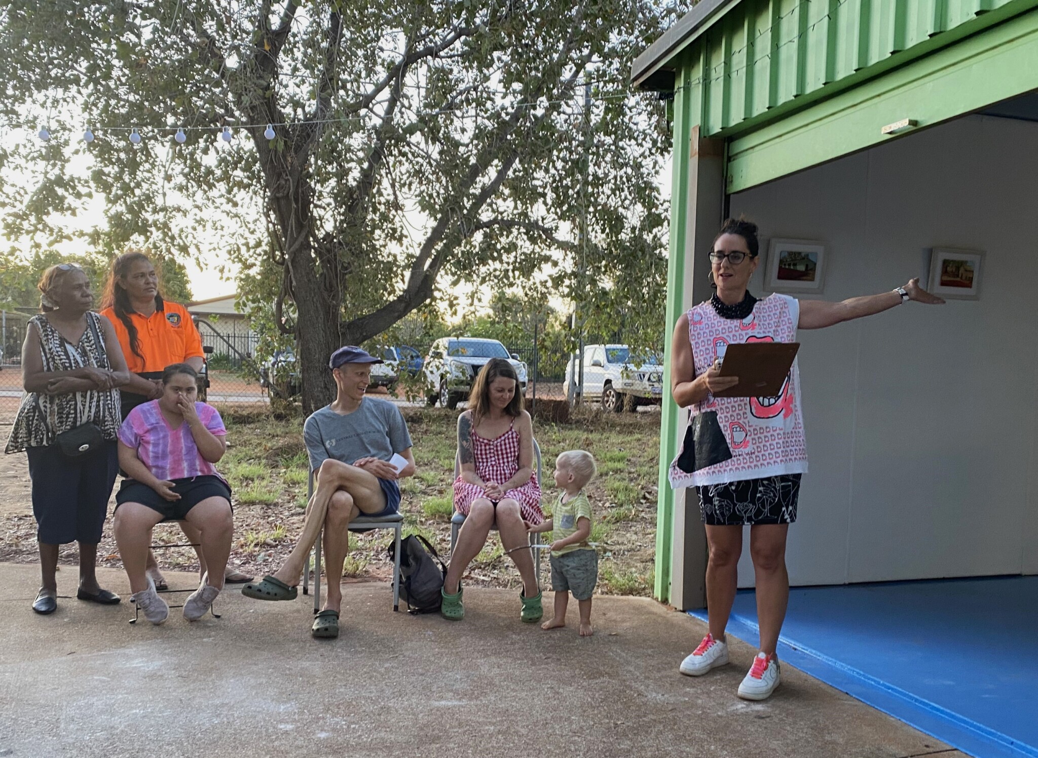 Five adults and a toddler sit and stand, watching as a woman officially opens the shed. She holds a clipboard in one hand and has her other arm outstretched.