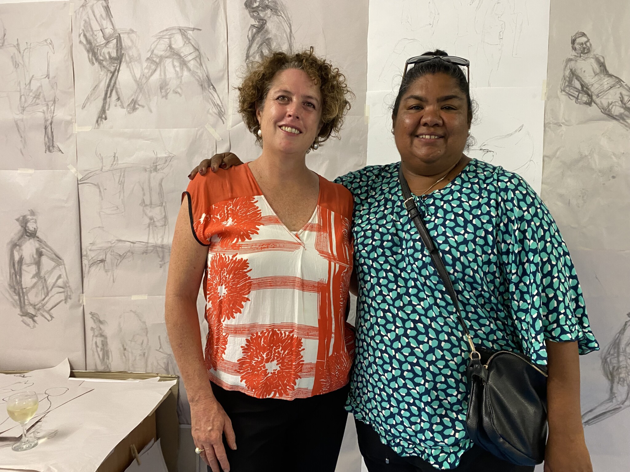 Two women stand smiling in front of taped up life drawing sketched on butchers paper.