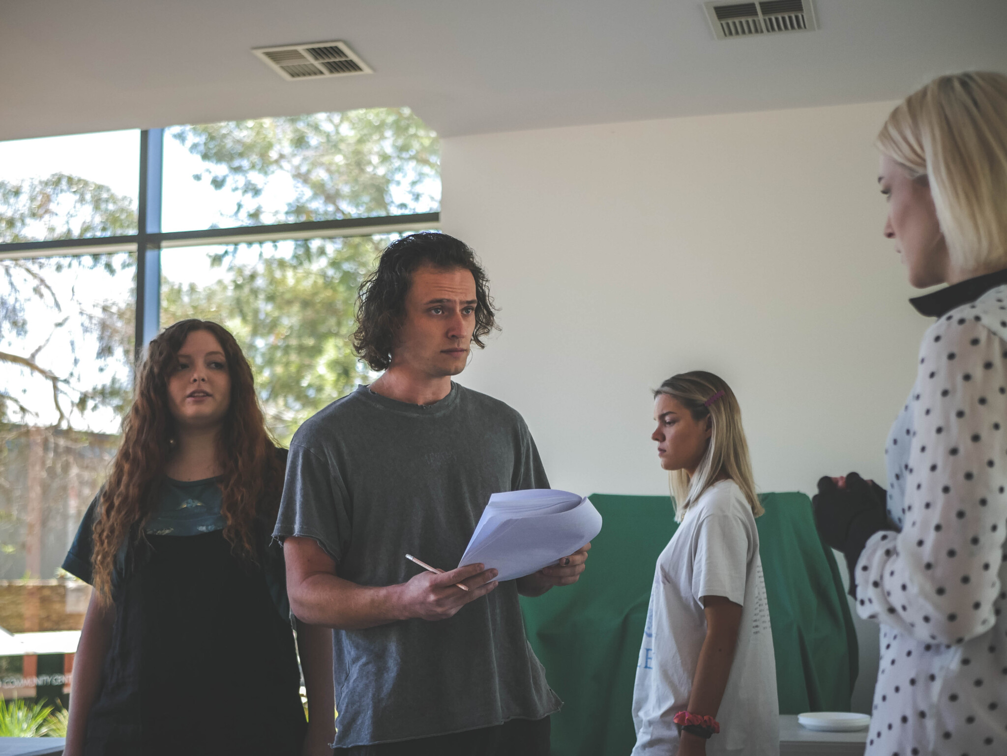 Four people indoors; one holds papers and pen, appearing to explain something. Large windows show trees, green fabric nearby.