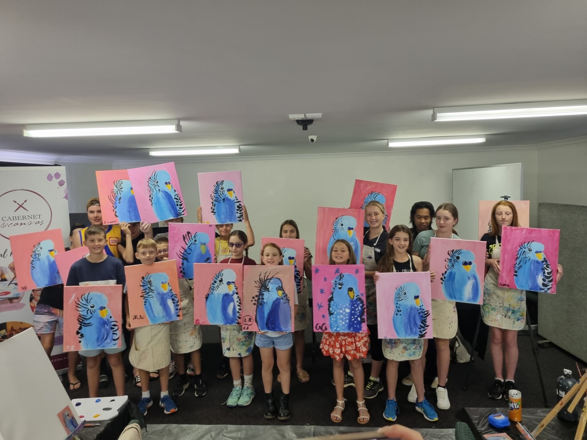 Seventeen children show off their blue budgie paintings from their workshop.