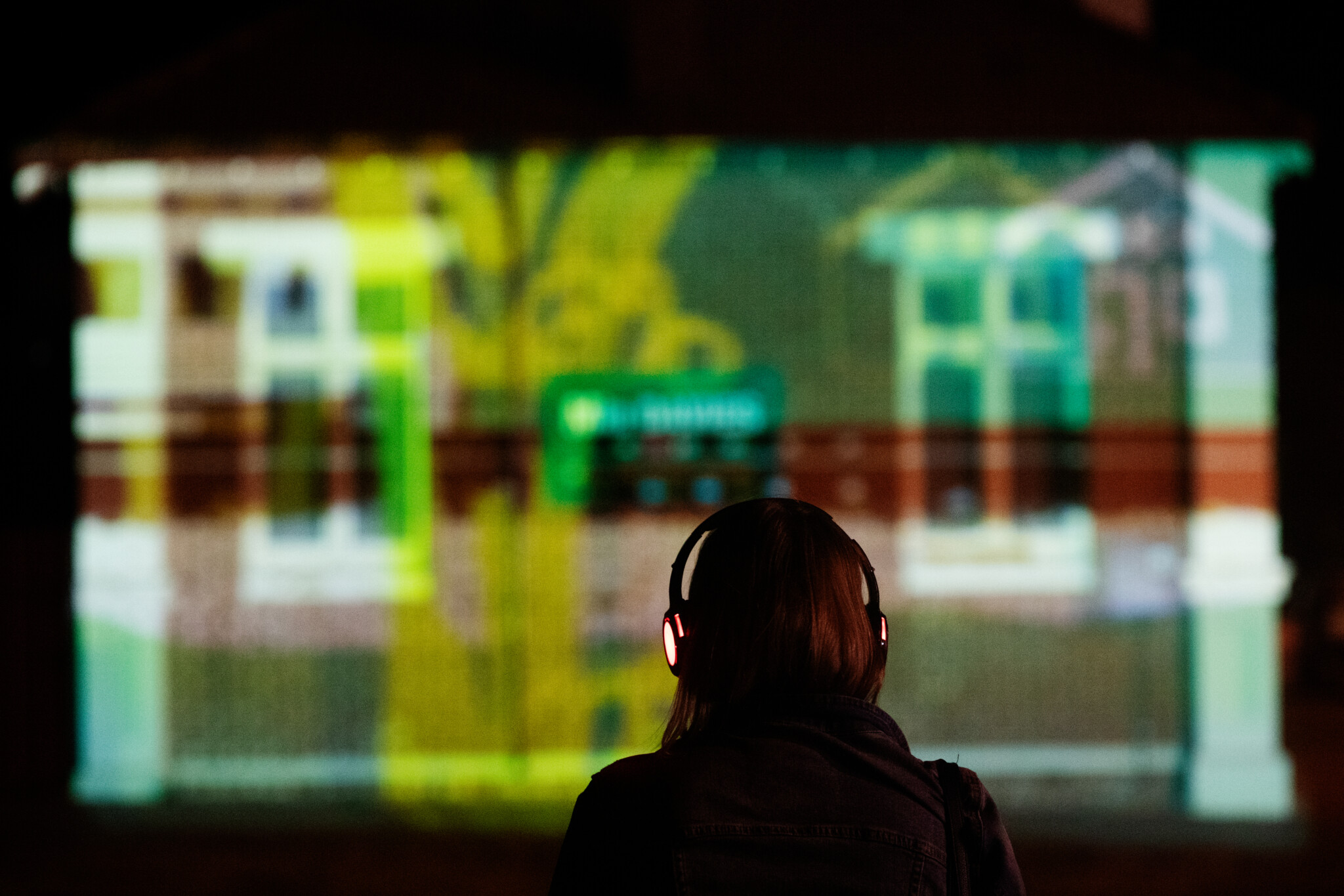A person wearing headphones has their back turned to the camera, they are looking at an abstract light projection.