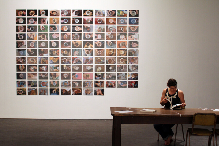 Person seated at table reading a book; wall behind features grid of small photos showing top views of coffee cups.