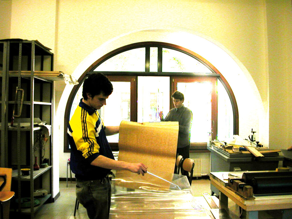 Francesco as a teenager in a workshop. He is holding a large sheet of paper which he is guiding into a large sheet of plastic. He is looking down and concentrating.
