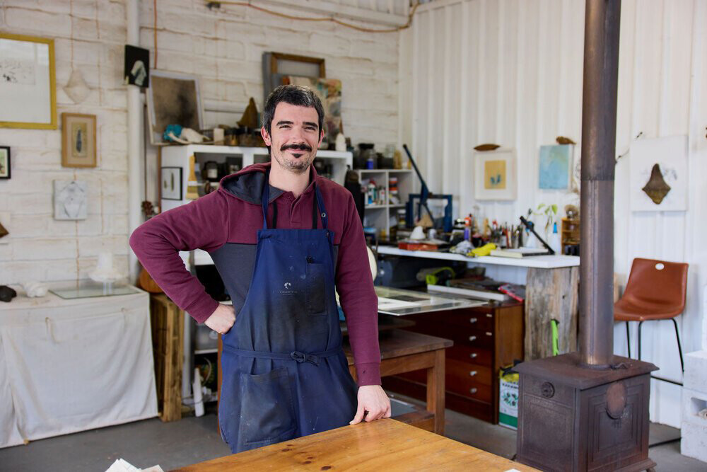 Francesco standing in his printmaking studio. He is smiling, and wearing a maroon hooded jumper and a dark blue apron. His workshop has lots of artwork hung up on the wall, and different printmaking equipment.