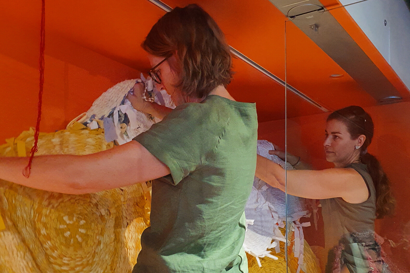 Two people work on colorful textile installation in orange-walled space; one wears glasses and green shirt, other has face obscured.