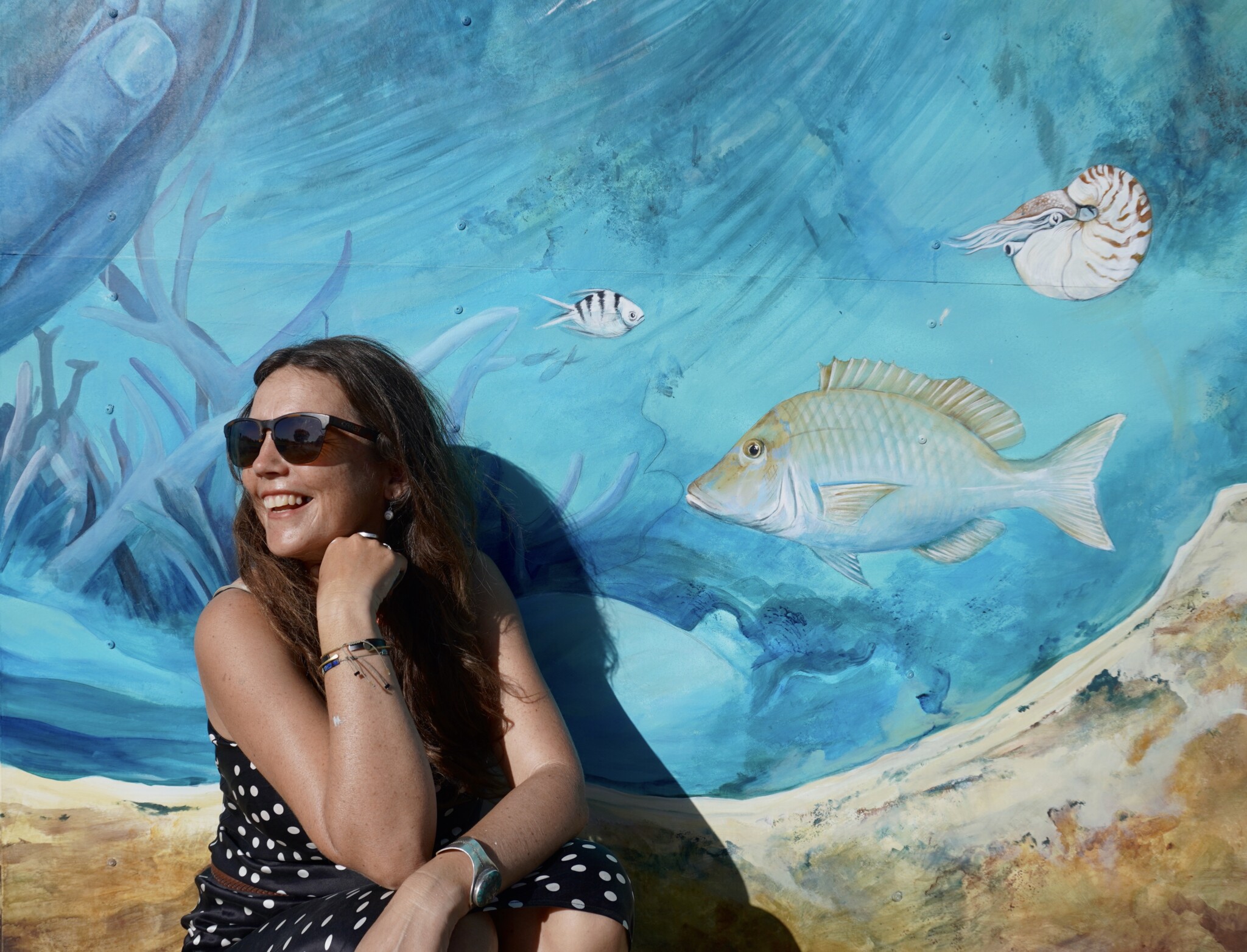 Woman crouches next to ocean mural while smiling off camera