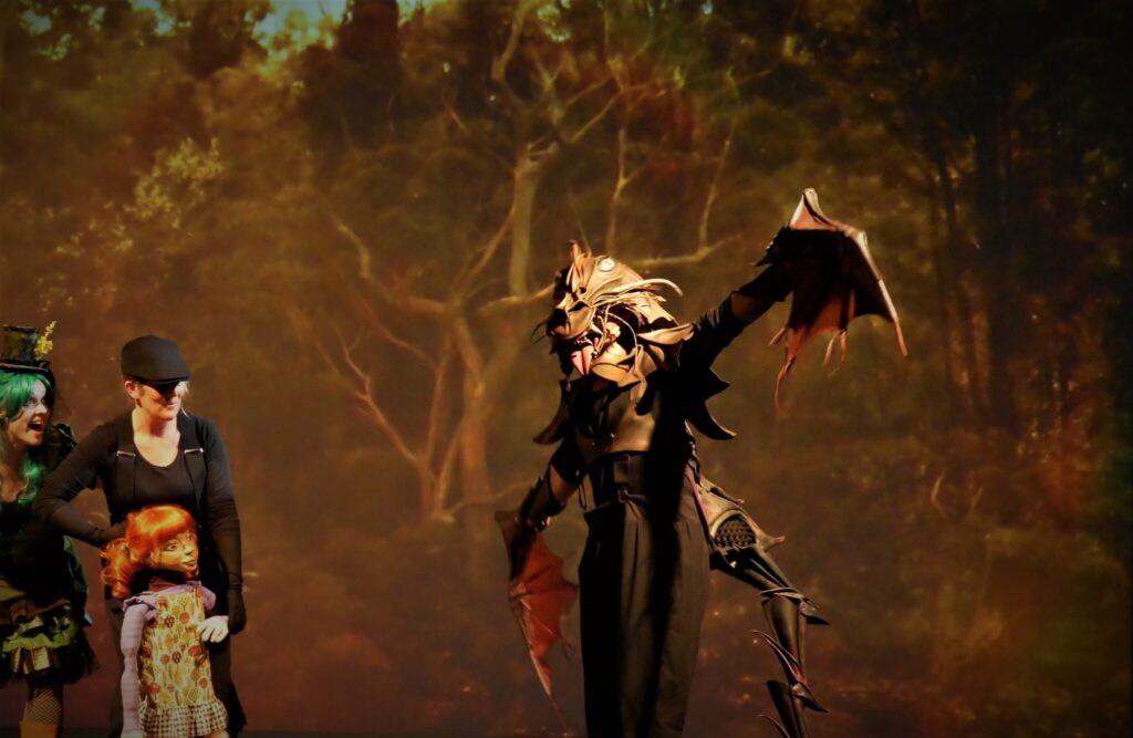 Person in elaborate dragon costume with wings stands on stage; adult and child in costume nearby, forest backdrop behind them.