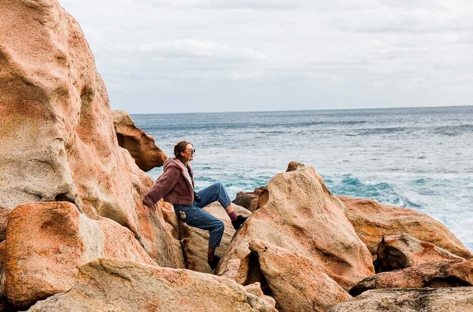 Woman sitting against rocks, wearing pink fluffy jacket and jeans, the ocean is in the background.