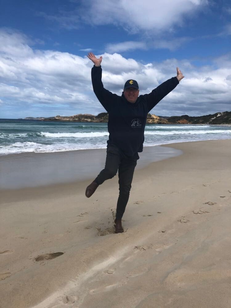 Martin Tunley, wearing a hat and jumper on the beach, doing a dynamic pose
