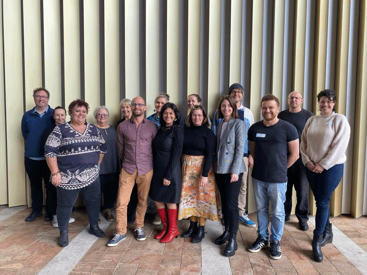 A group of 15 people stand in front of a building wall, smiling at the camera