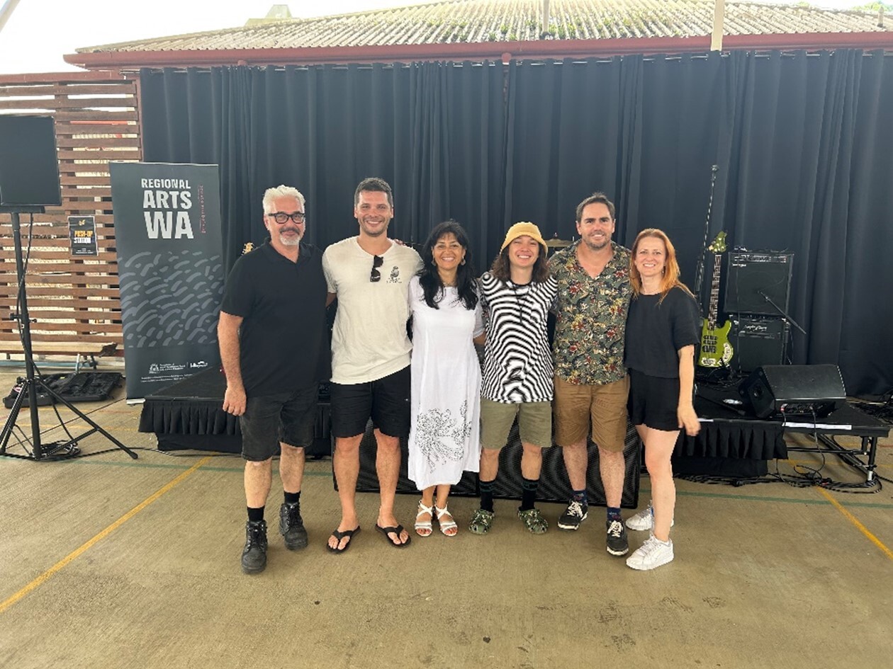 Six people pose in front of black curtain with "REGIONAL ARTS WA" banner; guitar and amps visible, outdoor concrete floor setting.