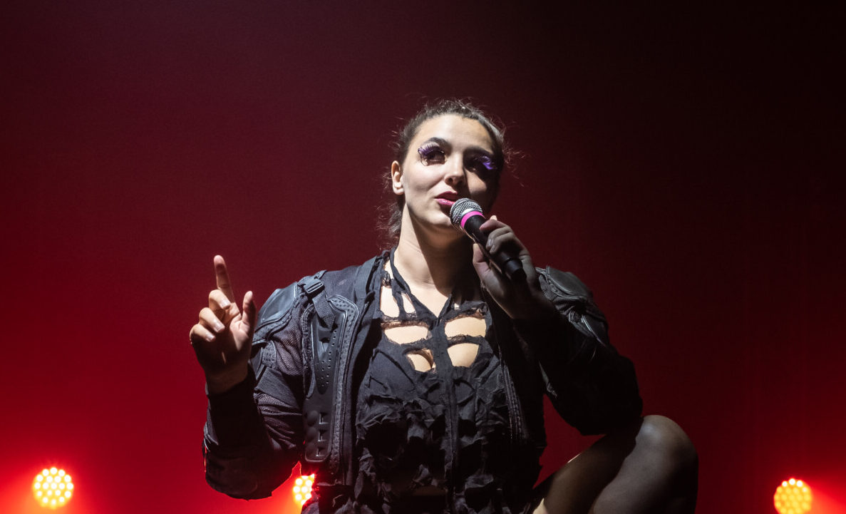 A woman stands on stage with a microphone, wearing dark ornate clothing, red lighting illuminates the background.