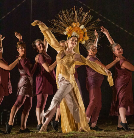 Central dancer in gold costume with floral headpiece performs on stage, surrounded by dancers in maroon striking expressive poses.