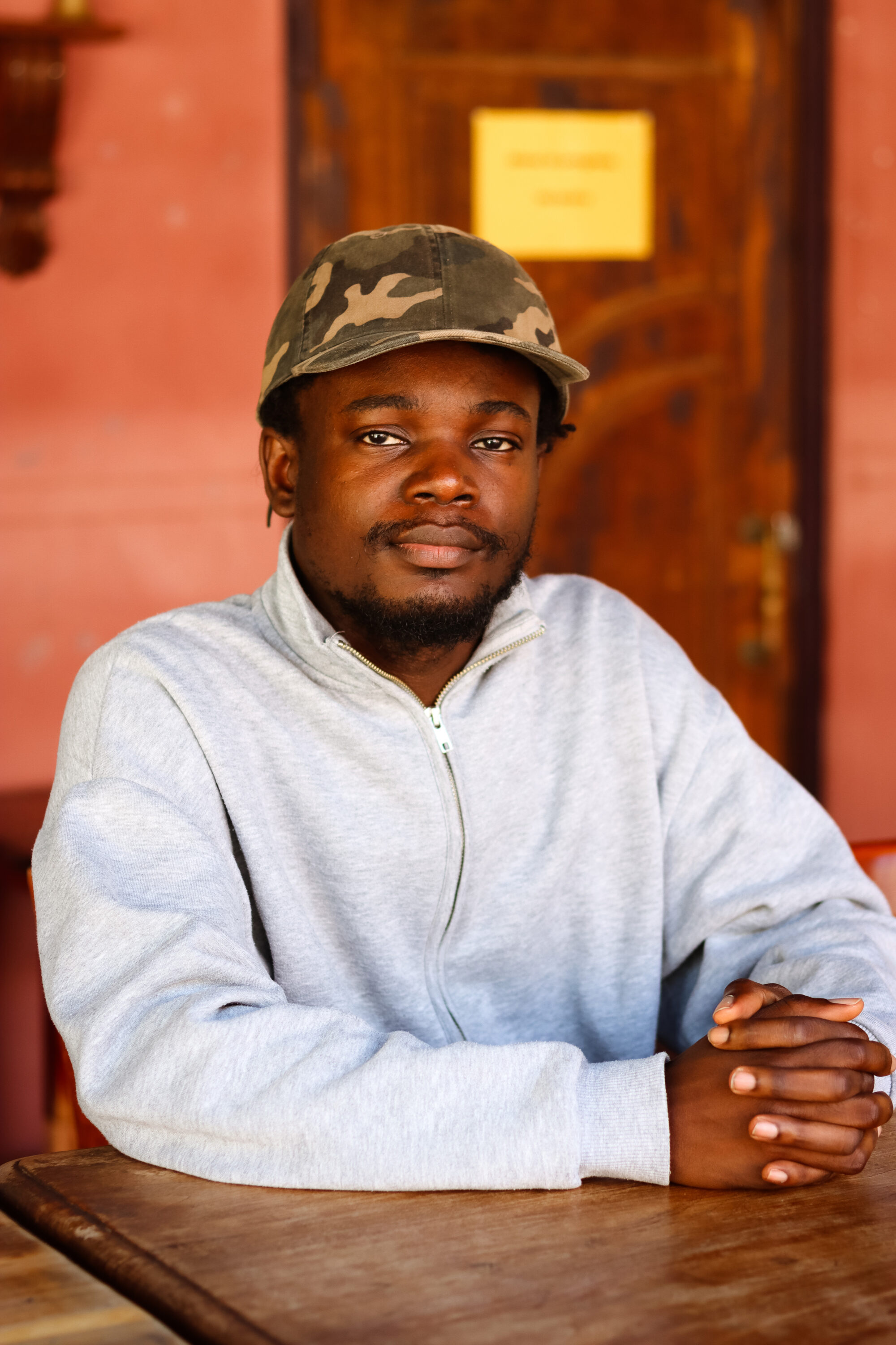 Headshot of Mohammed 'Ayo' Busari wearing a camo hat and a light grey jumper